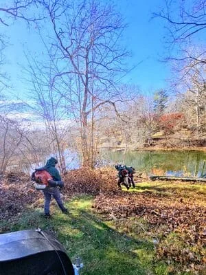 Crew performing leaf and debris removal on waterfront property with bare trees and water visible